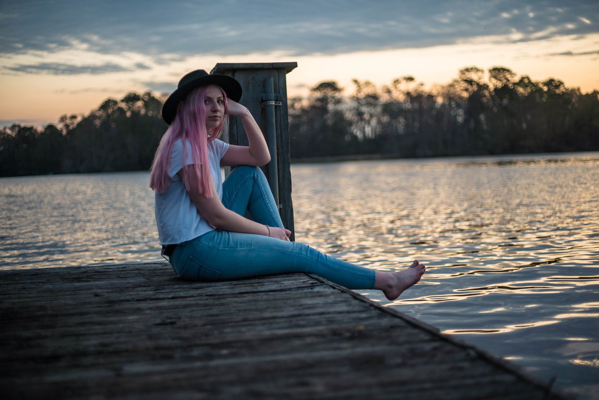 Young woman with ah hat and pink hair sitting on a dock byt the water with her leg extended