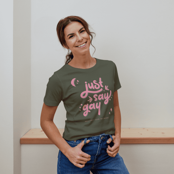 woman with wavy hair in a ponytail wearing a fitted olive green colored t-shirt with pink script words that say "Just Say Gay"